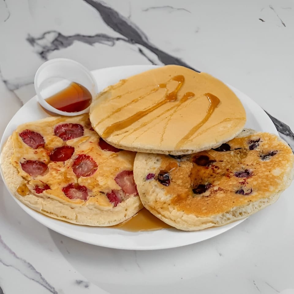Three pancakes, one plain, one strawberry, and one blueberry, drizzled with syrup on a white plate.